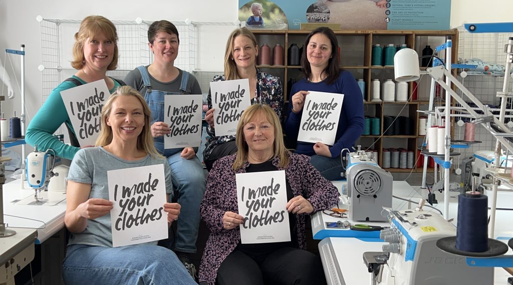 A group of people in a sewing factory, holding up signs that say 'I make your clothes'.