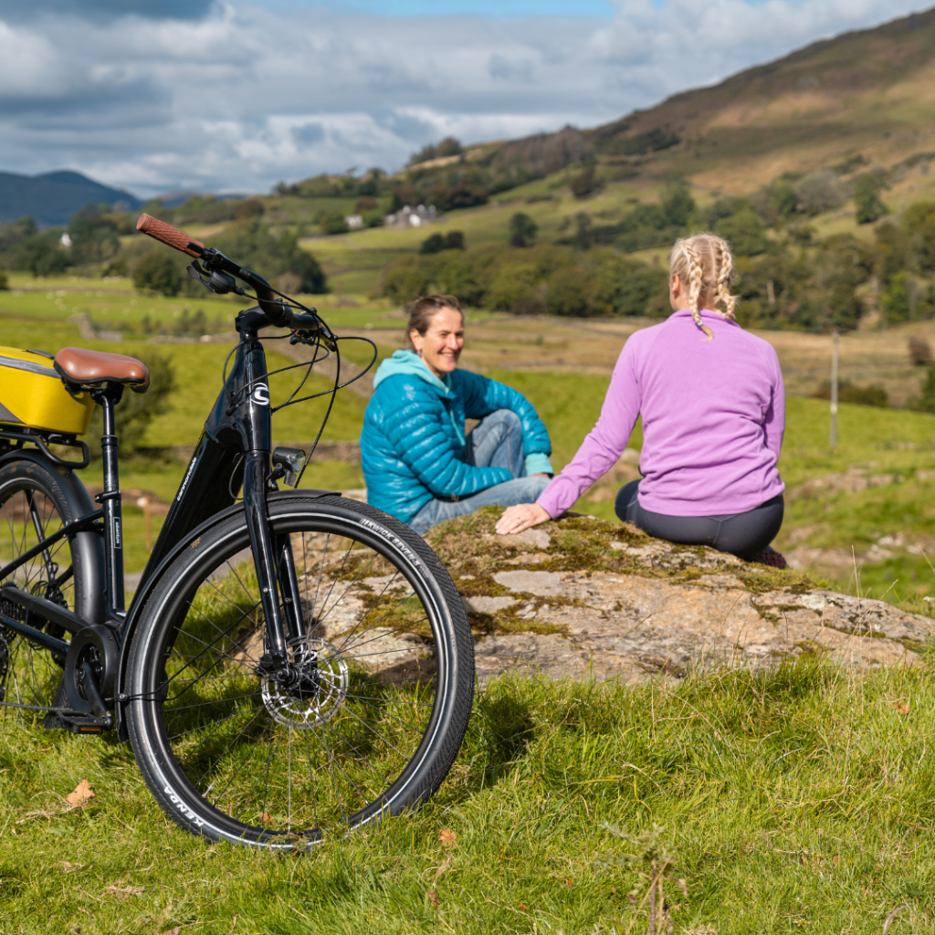 Two people sitting on a log in a field, with a hill in the background and an ebike in the foreground.