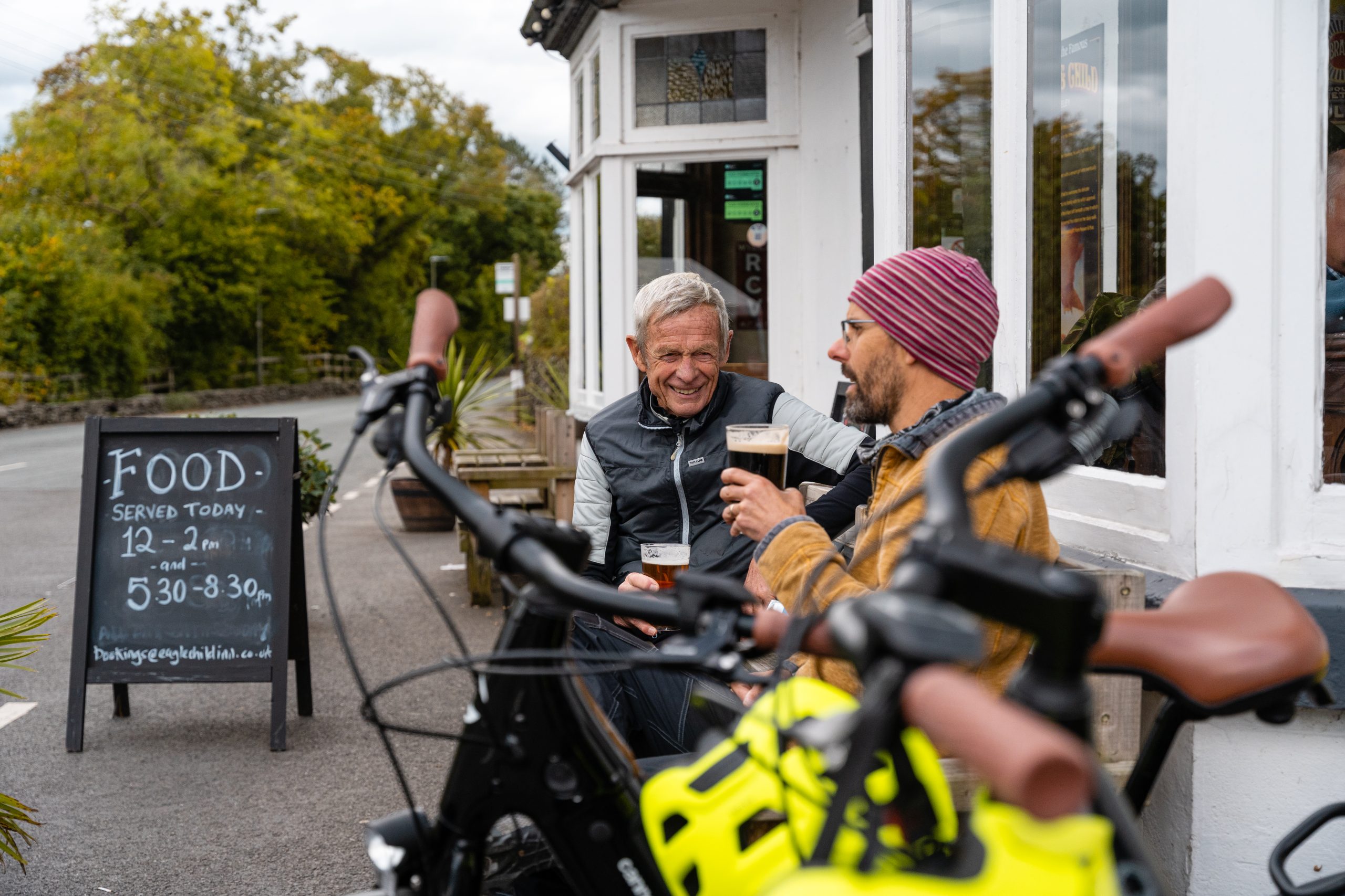 Two people sitting outside the Eagle and Child Pub in Staveley, with ebikes standing nearby.