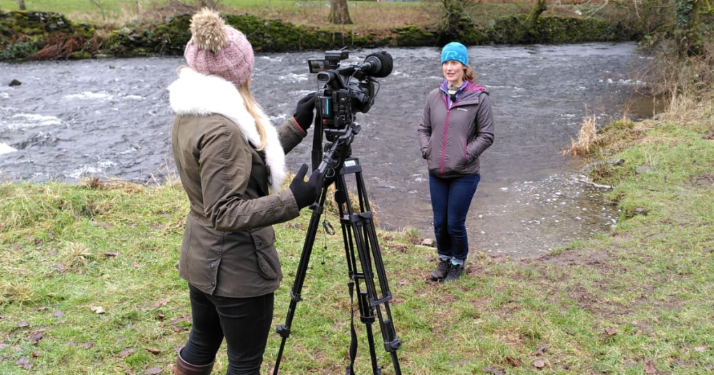 A person with a camera on a tripod filming another person talking about the River Kent, which they're standing in front of on a grassy bank. 