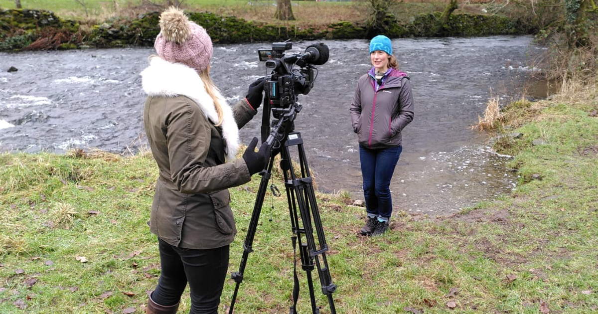 A person with a camera on a tripod filming another person talking about the River Kent, which they're standing in front of on a grassy bank.