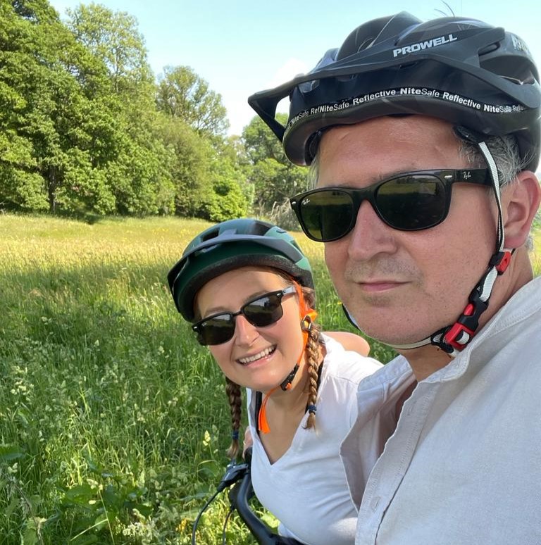 Two people with ebikes in front of a summer meadow and trees, wearing helmets and sunglases.