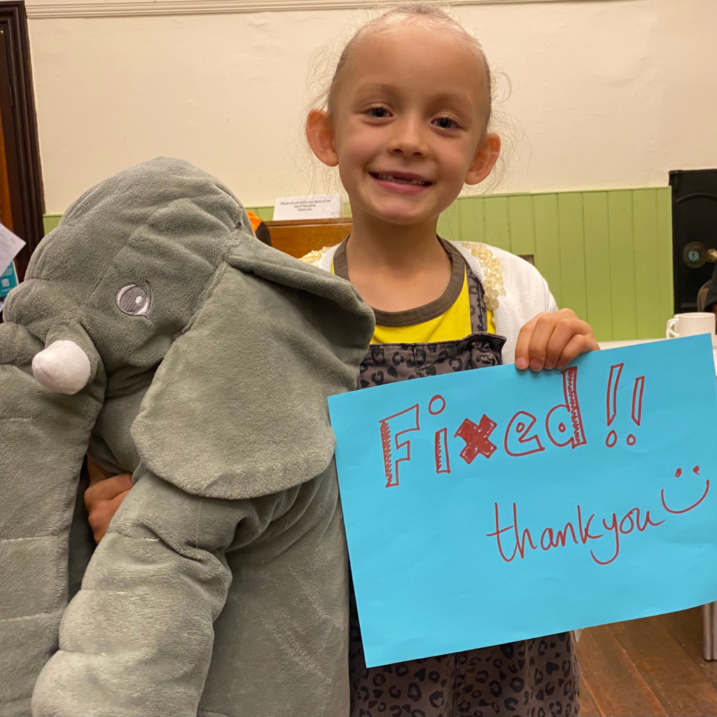 A young girl holding an elephant cuddly toy and a sign that says 'fixed!'