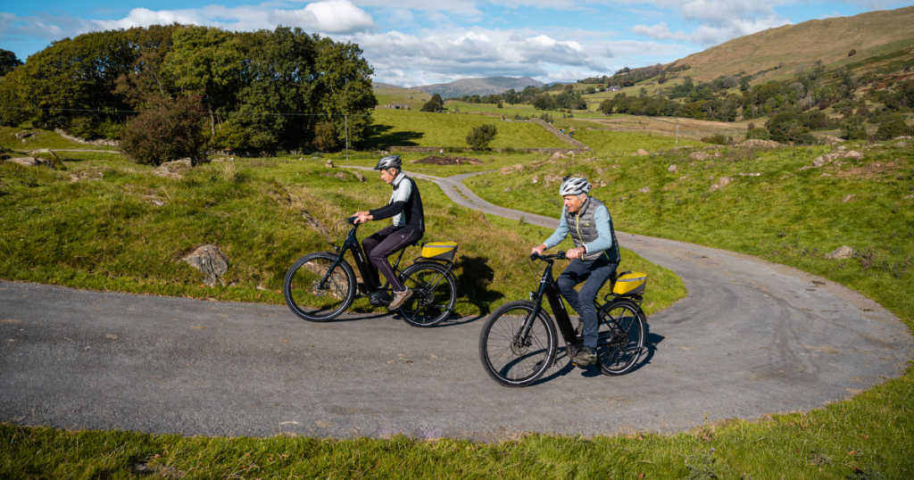 A shot of two people cycling on ebikes up a slight hill on a country road.