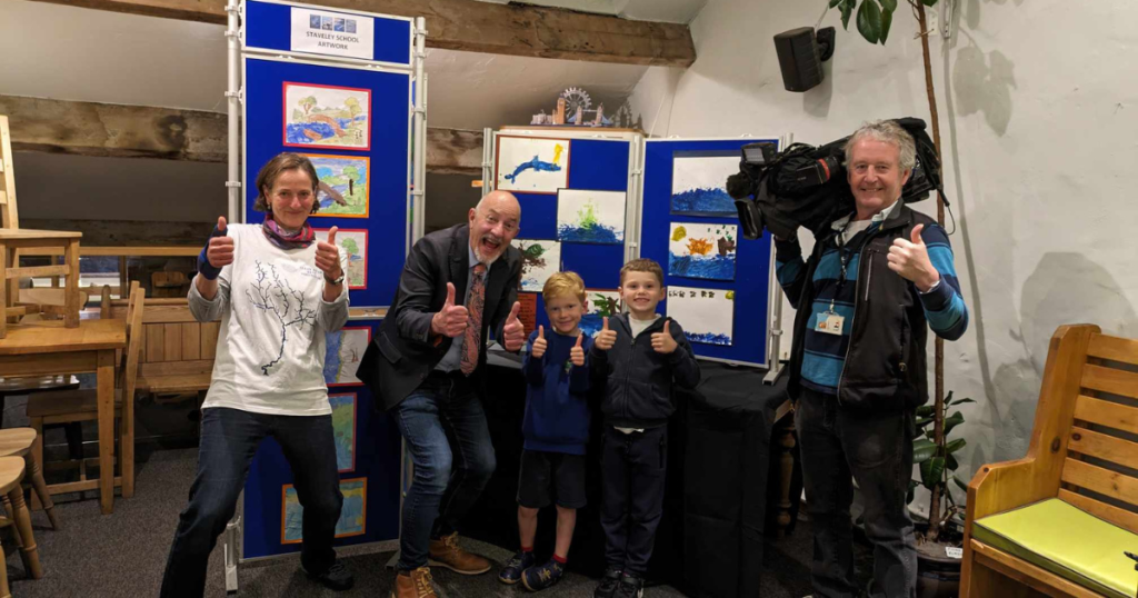 A group of adults and children showing a thumbs up to the camera while taking part in a media interview at the SENS Along the River Kent art exhibition. 