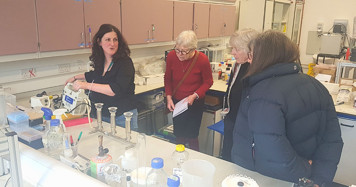 A group of people standing in a science lab at the University of Sheffield with scientific equipment in front of them, learning about microbial source tracking. 