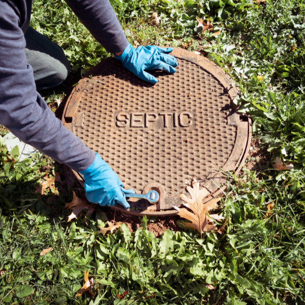 An overhead view of a septic tank cover being removed by a person wearing blue plastic gloves.