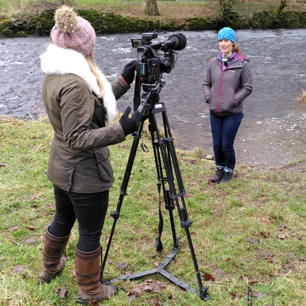 Two people, one behind a camera, filming at the side of the river Kent in Staveley.