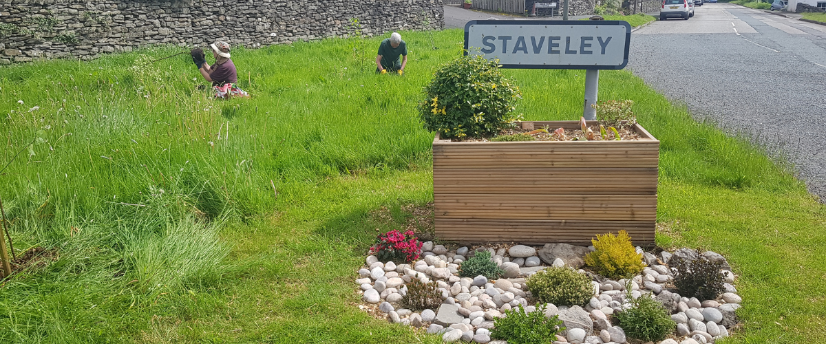 The entrance to Staveley from the Windermere side, showing a sign with a box planter and plants underneath. 